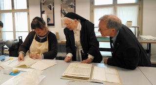 In het restauratieatelier geeft een behoudsmedewerker uitleg over de conservering van een archiefstuk. Twee bezoekers kijken toe.