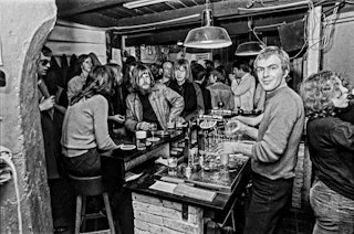 Een barman kijkt in de lens, voor de bar staat het vol met mensen.