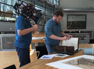 Links een cameraman met een camera op zijn schouder en rechts staat een man die bladert in een archiefstuk.