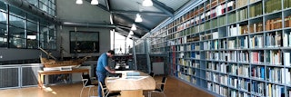Een grote ruimte met rechts een oneindige wand met boeken en centraal een tafel met archiefstukken, waarover een man zich buigt.