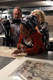 Een cameraman filmt een vrouw die een kaart bekijkt en wijst naar een plek op de kaart.