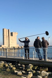 Een filmploeg staat op een loopbrug, op de achtergrond is een pijler in het water te zien.