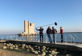 Een filmploeg staat op een loopbrug, op de achtergrond is een pijler in het water te zien.