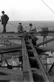 Staand op stalen balken hoog boven de grond kijken twee mannen recht de camera in. Rechs van hun werken vier arbeiders staand en zittend op de balken. In de verte is de abdijtoren van Middelburg te zien.