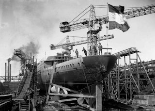 Een schip zonder opbouw, met een wapperende Nederlandse vlag vanaf de achtersteven, ligt op een scheepshelling.