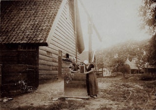 Twee vrouwen staan bij een waterput bij de achterkant van een schuur.
