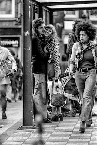 Een man en een vrouw omhelzen elkaar in de abri van een tram.