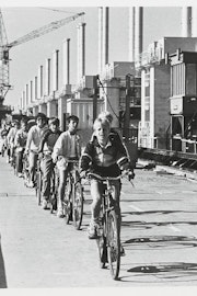 Scholieren fietsen achter elkaar aan over de Oosterscheldekering, op de achtergrond een bouwkraan..