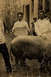 Drie mannen met een groot varken staan in een straat.