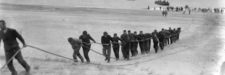 Zwart-witfoto van mannen die kabel uit de zee aan land trekken.