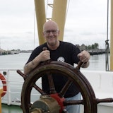 Een glimlachende man staat achter een stuurwiel aan dek van een schip.