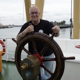 Een glimlachende man staat achter een stuurwiel op het dek van een schip.