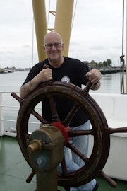 Een glimlachende man staat achter een stuurwiel op het dek van een schip.