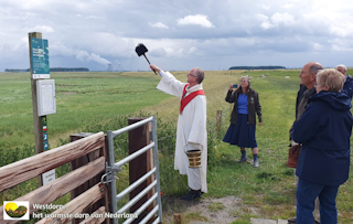 Hek met mensen ervoor. In het midden een priester die een zegen uitspreekt. Op de achtergrond schorren in Saeftinghe