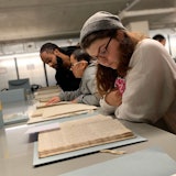 Twee studentes en hun docent buigen zich over archiefstukken die op metalen archiefkasten liggen. Ze bekijken handschriften.