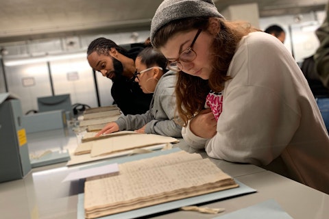 Twee studentes en hun docent buigen zich over archiefstukken die op metalen archiefkasten liggen. Ze bekijken handschriften.