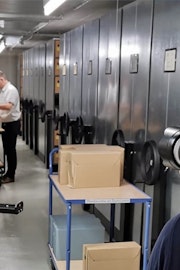 Een archiefmedewerker opent een archiefdoos die op een archiefkar in een gang in het depot staat.