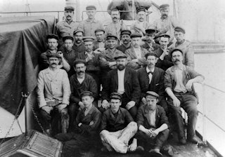 Een groep mannen poseert zittend en staand aan het dek van een schip.