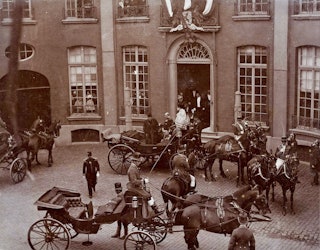 Gezicht op het halfronde plein voor een statig huis. Een man en vrouw nemen plaats in een open koets met vier ingespannen paarden.