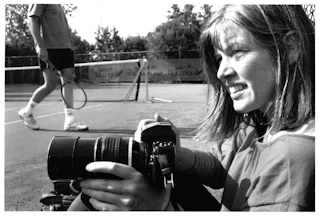 Een vrouw met fototoestel in haar handen zit gehurkt naast een tennisbaan.