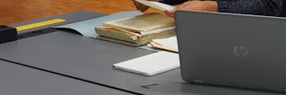 Op een tafel is een laptop opengeklapt, erachter zijn iemands handen te zien. Op de tafel liggen stapeltjes papieren.