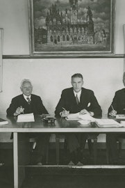 zwart-witfoto van vier mannen die zitten achter een tafel en recht de camera in kijken.