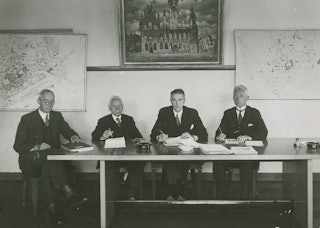 zwart-witfoto van vier mannen die zitten achter een tafel en recht de camera in kijken.